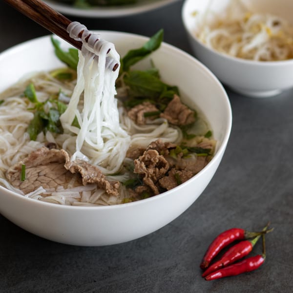 Chopsticks lifting noodles from a bowl of Pho City pho with beef, herbs, and red chilies.