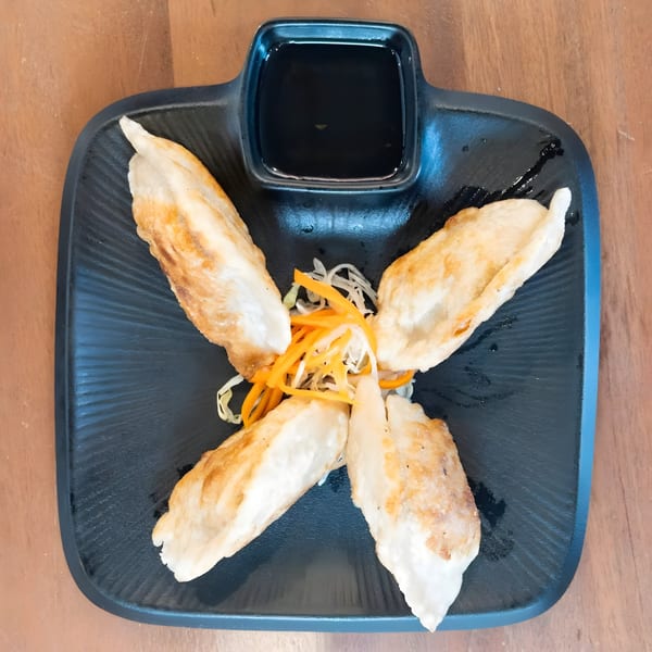 Four golden-brown fried dumplings with shredded carrots and cabbage, served with soy sauce on a black plate.