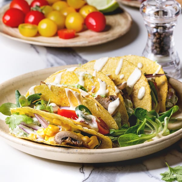 Three hard-shell tacos filled with mushrooms, corn, tomatoes, and greens, drizzled with white sauce.