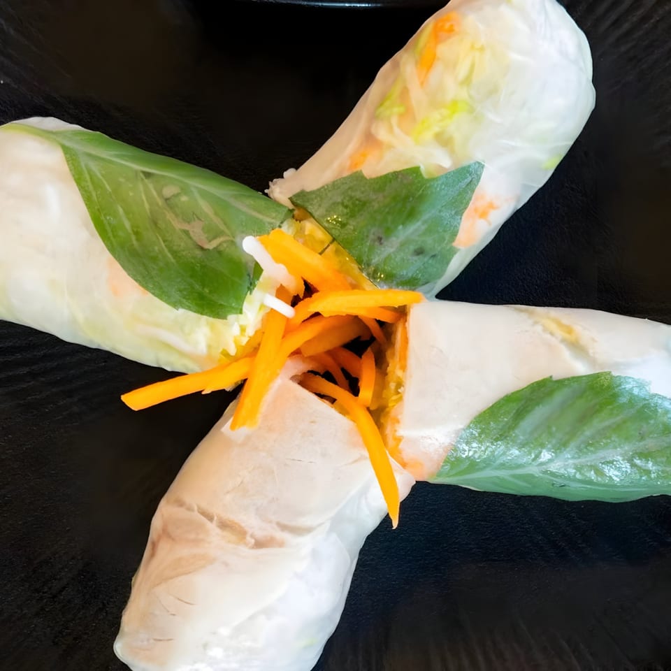 Two fresh Vietnamese spring rolls, filled with carrots and greens, crossed on a dark plate.